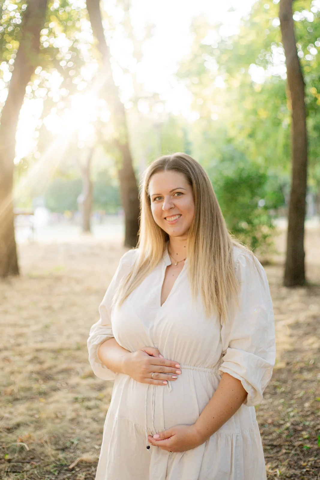 Kismama világos ruhában, fák között.